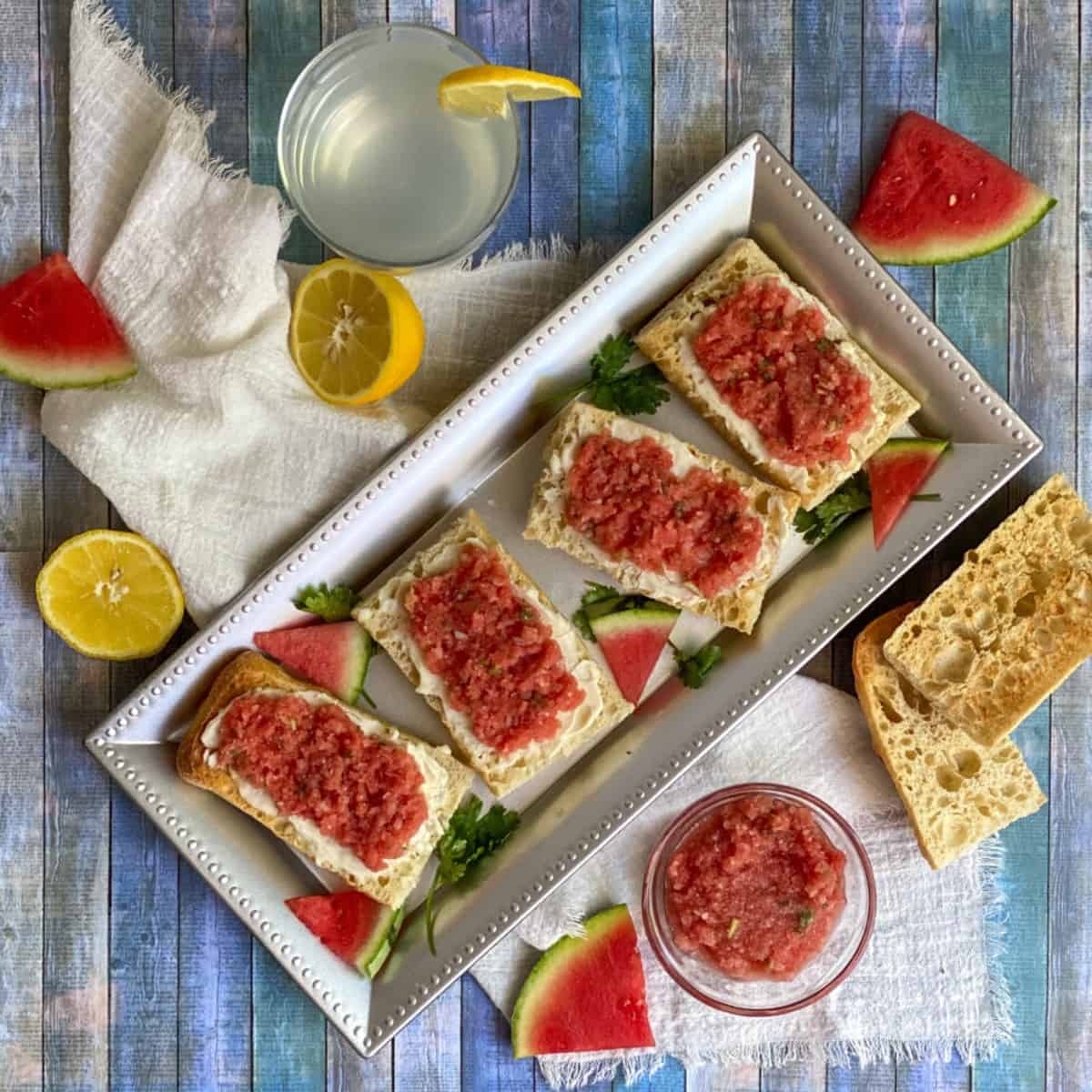 A rectangular tray with a slice of bread in the center. The bread is topped with a mixture of diced red and yellow crimson watermelon, chopped fresh mint, and a white feta cheese.