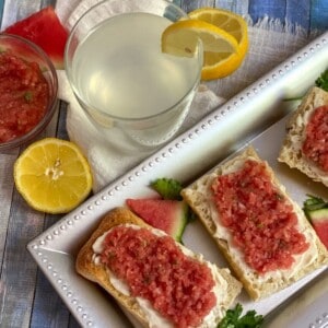A close-up of two slices of breakfast bruschetta topped with watermelon slices and cream cheese on a wooden tray. There are also a few small sprigs of fresh mint scattered around the plate.
