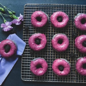 a close-up of several blackberry donuts glazed with a dark purple colored frosting. The donuts are round with a hole in the middle and are sitting on a wire rack.