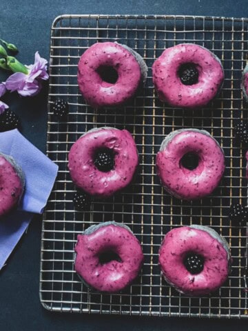 a close-up of several blackberry donuts glazed with a dark purple colored frosting. The donuts are round with a hole in the middle and are sitting on a wire rack.