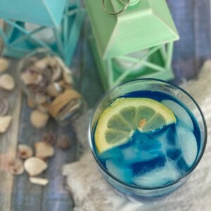 A tall glass filled with bright blue curacao lemonade colored lemonade with ice cubes. A slice of lemon rests on the rim of the glass.