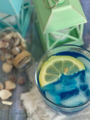 A tall glass filled with bright blue curacao lemonade colored lemonade with ice cubes. A slice of lemon rests on the rim of the glass.