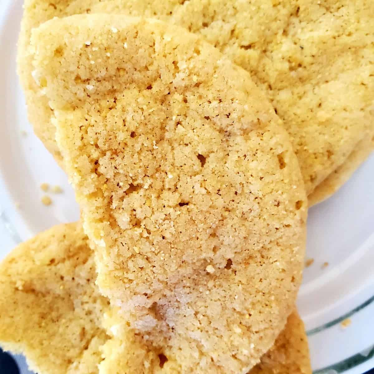 A close-up of a single corn cookie on a white plate. The cookie is round and golden brown with visible yellow corn kernels throughout. It has a slightly cracked and bumpy surface, and there are crumbs on the plate around the cookie.