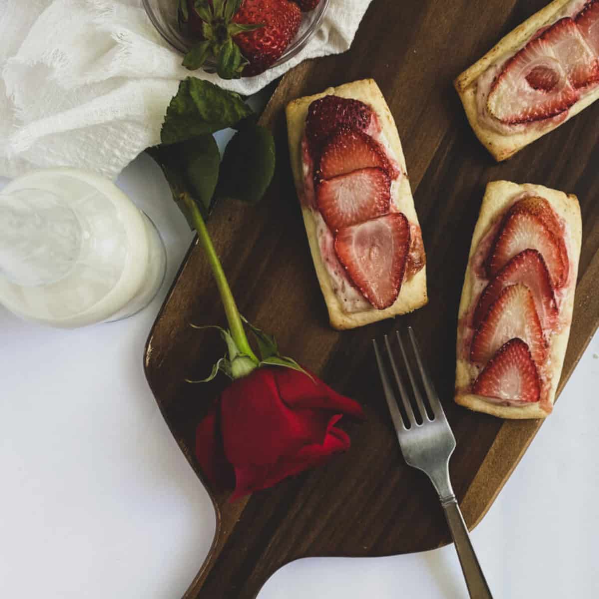 strawberries and cream puff pastry with sliced strawberries on a wooden cutting board with a fork and a rose