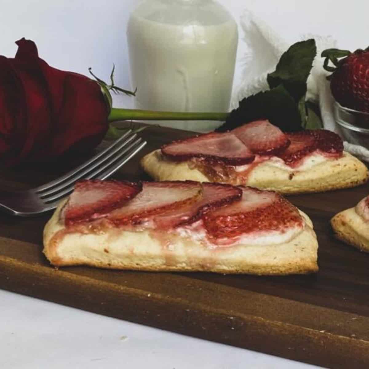 Two slices of strawberry gluten free puff pastry on a wooden cutting board. Each slice has a golden brown crust topped with sliced strawberries and cream cheese frosting.