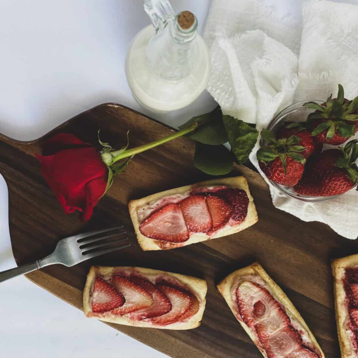 A close-up of a rectangular puff pastry turnover filled with cream cheese and sliced strawberries. The pastry is golden brown and flaky, with a dusting of powdered sugar on top. The strawberries are bright red and juicy.