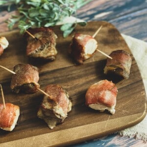 A close-up of several bite-sized pieces of bacon wrapped steak bites on a white plate. The steak and bacon are cooked and golden brown. There is a sprig of fresh rosemary on the plate for garnish.