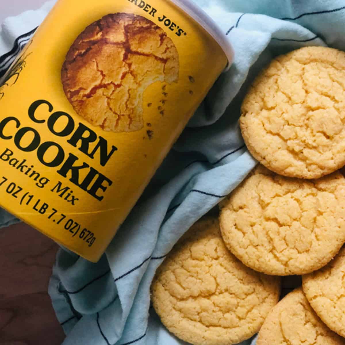 A can of Trader Joe's Corn Cookie Mix next to a basket of golden brown cookies on a white towel. The text on the can says "TRADER JOE'S" in large white letters, with "CORN COOKIE" and "Baking Mix" written below it. The weight of the mix is listed as "7 OZ (1LB 7.7OZ) 672g"