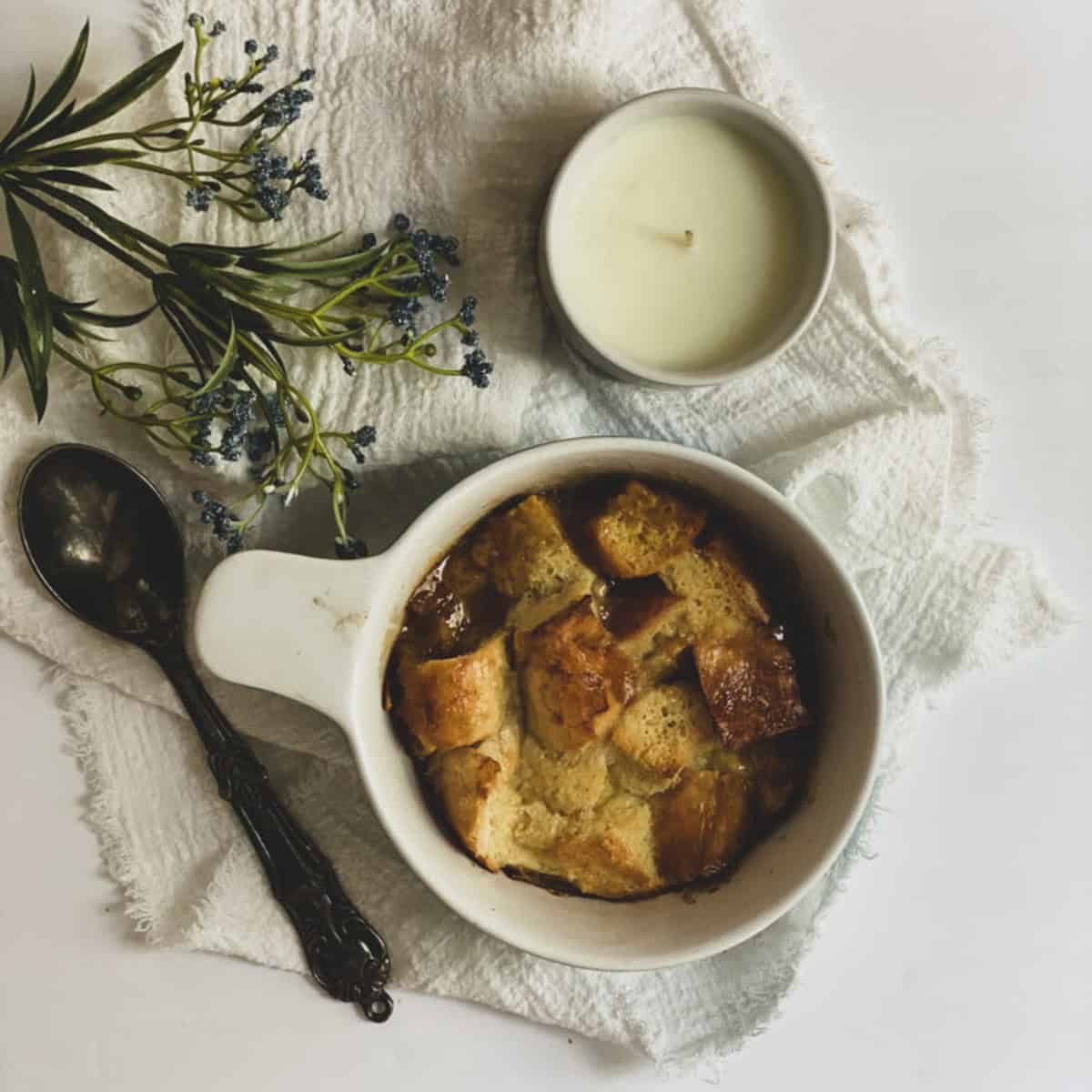 A bowl of bread pudding on a white napkin. The bread pudding is light brown and sits in a white bowl.
A white spoon rests on the edge of the bowl.
To the right of the bowl is a white mug filled with milk.
To the left of the bowl is a lit white candle in a glass holder. This is a breakfast to feed a large crowd