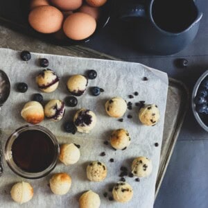 pancake bites scattered with blueberries and chocolate chips. There are also some apple slices on the platter. While some of the pancakes are golden brown, others are a darker brown. a cup of maple syrup is on the sheet with the pancakes.