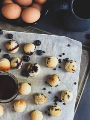 pancake bites scattered with blueberries and chocolate chips. There are also some apple slices on the platter. While some of the pancakes are golden brown, others are a darker brown. a cup of maple syrup is on the sheet with the pancakes.