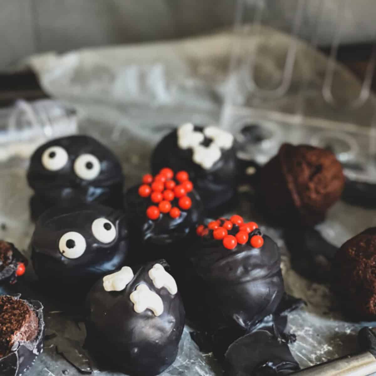 A close-up of a rectangular tray filled with round chocolate truffles. The truffles are decorated for Halloween with colorful sprinkles and some have googly eyes stuck on them. A small silver server sits on the edge of the tray.