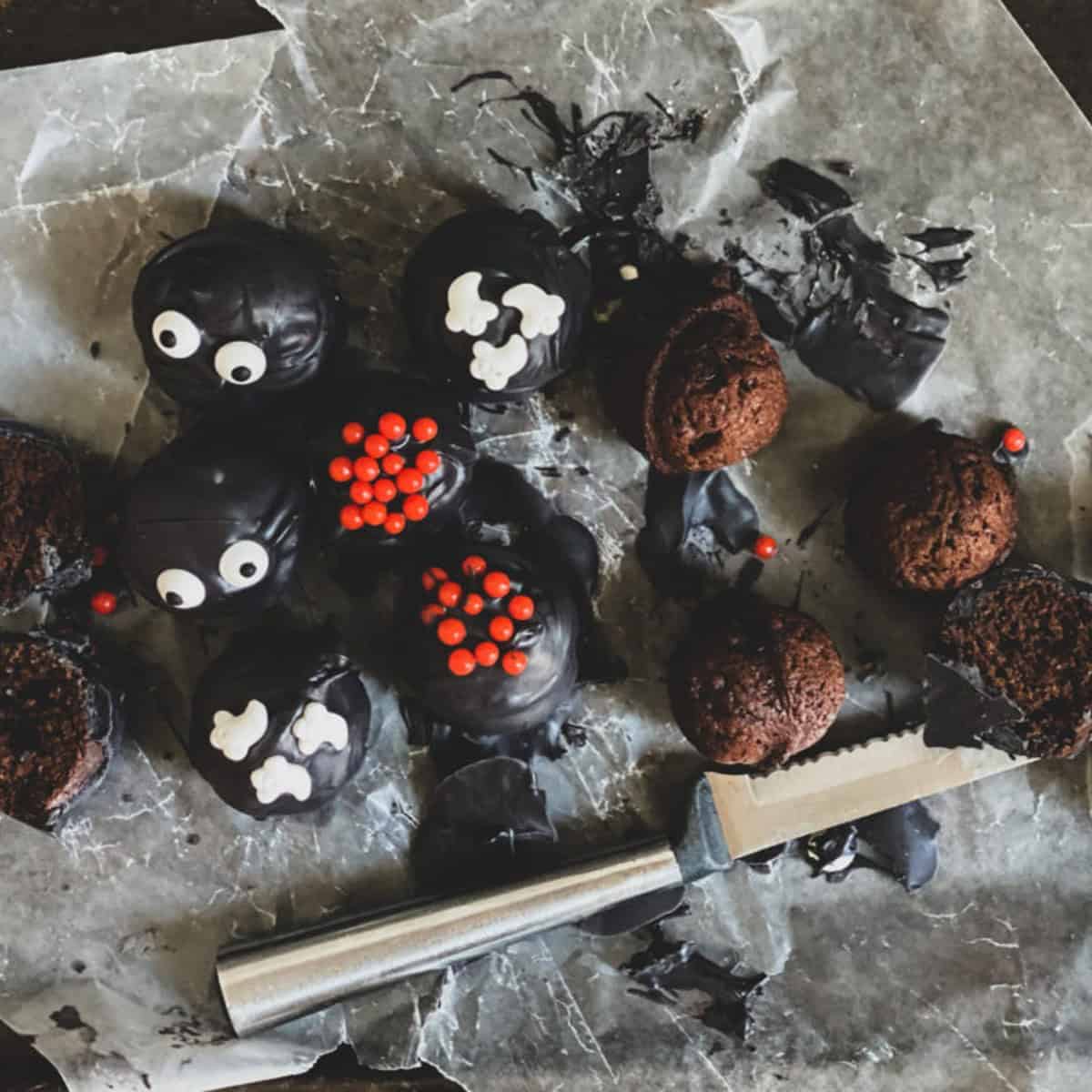 A close-up of a plate filled with chocolate cake ball treats. The treats are round and dusted with colorful sprinkles. Some treats have googly eyes added for a spooky look. A small silver knife sits on the edge of the plate.