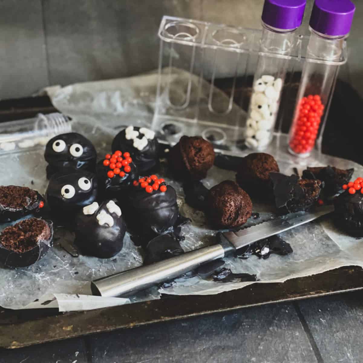 A tray of Halloween- truffles. The truffles are chocolate, round and dusted with colorful sprinkles. Some truffles have googly eyes added for a spooky look. A silver knife sits on the edge of the tray.