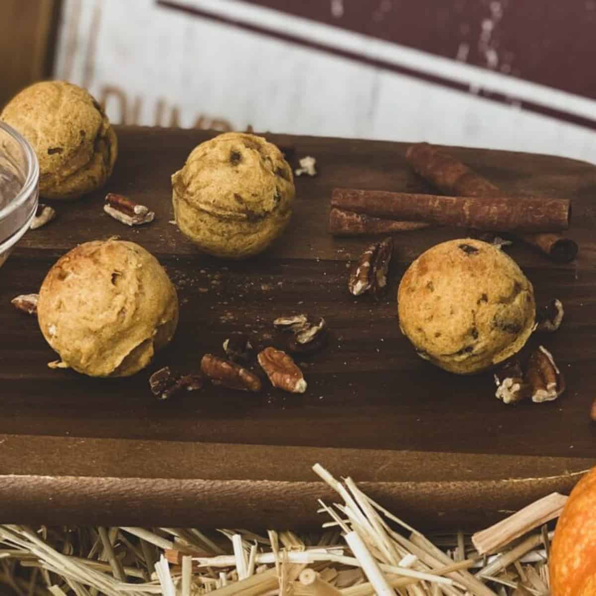 Four mini pumpkin pancakes with pecans arranged on a wooden board. The pancakes are golden brown and topped with chopped pecans. Cinnamon sticks and additional pecan pieces are scattered around the pancakes. A small bowl of syrup is blurred in the background.
