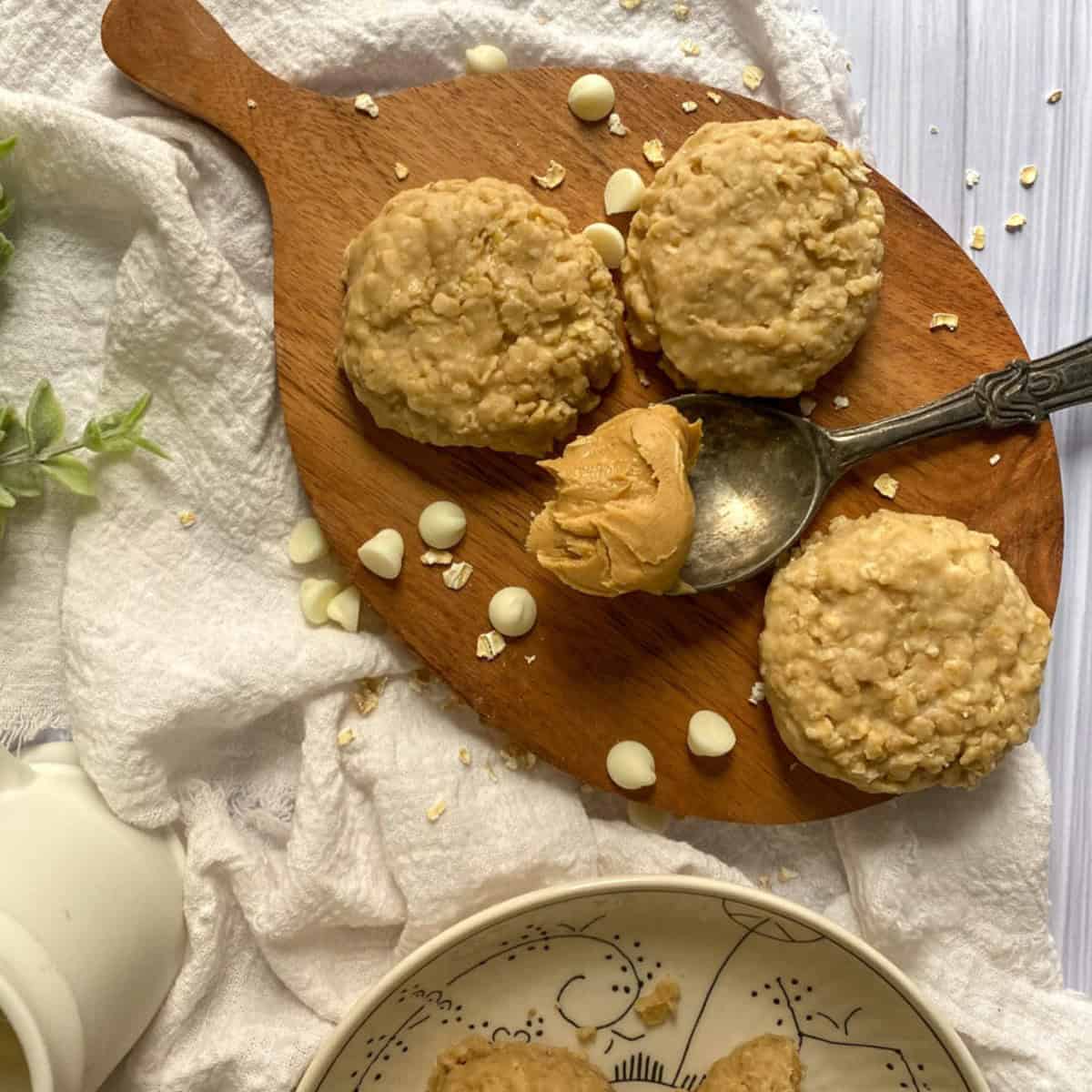 three peanut butter white chocolate cookies on a wooden board. The cookies are golden brown and appear to have a soft, chewy texture. There are also some white chocolate chips scattered around the cookies. In the background, there is a white cloth napkin, a silver spoon with peanut butter on it, and a white coffee cup.