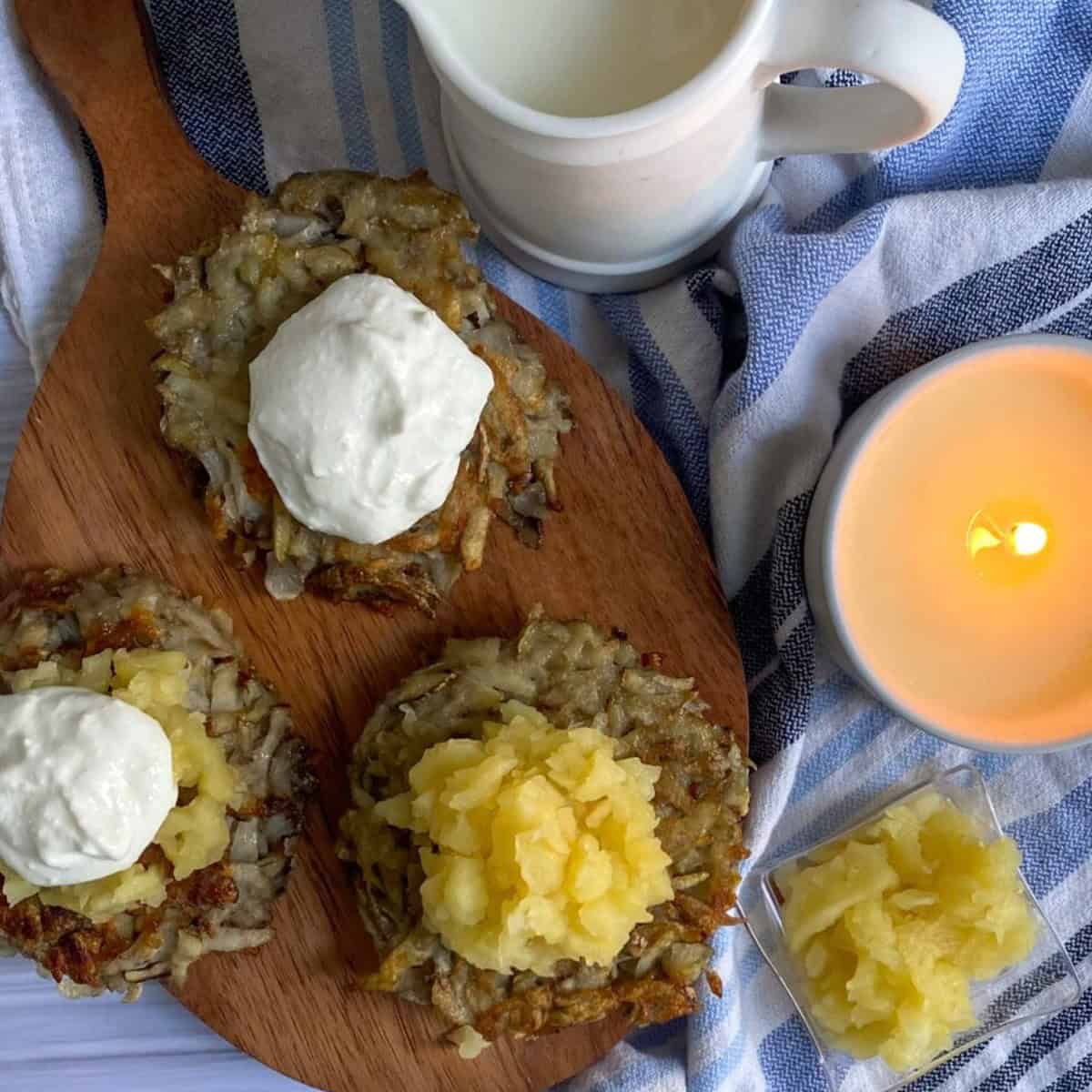 A close-up of a plate filled with three gluten-free potato latkes. The latkes are circular and golden brown with a slightly crispy appearance around the edges. They appear to be made from grated potato and have a flat surface with visible potato shreds. To the left of the latkes sits a small, round white ramekin filled with applesauce. To the right of the latkes rests a dollop of white sour cream. The latkes are all on a white plate with a patterned, blue rim.