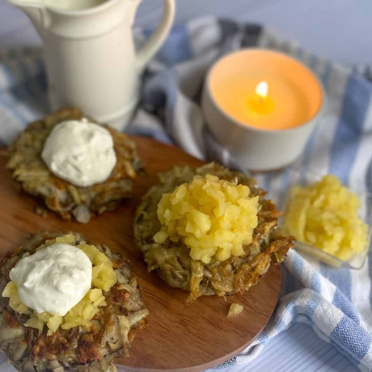 a photo of three potato latkes air fryer baked. They are arranged on a plate topped with applesauce and sour cream. A candle is lit for Hanukkah in the background. 