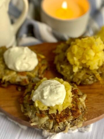 A round white plate filled with three gluten-free potato latkes. The latkes are golden brown and crispy around the edges, with a flat, grated potato surface. A small ramekin of applesauce sits to the left of the latkes, with a dollop of sour cream resting on the right.