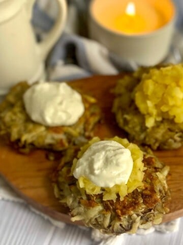 A close-up photo of three gluten-free potato latkes on a wooden cutting board. The latkes are golden brown and crispy-looking, with a slightly fluffy texture. They are topped with a dollop of sour cream and a spoonful of applesauce. The background is slightly blurred, but there is a hint of a kitchen counter in the background.