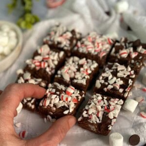 The image shows a plate of peppermint fudge recipe with marshmallows and sweetened condensed milk cut into squares. The fudge is dark brown and is topped with crushed candy canes. A hand is reaching for one of the squares. There are also marshmallows and chocolate chips scattered around the plate. The image looks delicious and festive, perfect for the holiday season.
