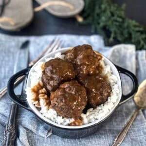 A bowl of white rice topped with four meatball halves and a rich, brown gravy. The bowl rests on a blue and white patterned cloth with a fork and spoon in the background.
