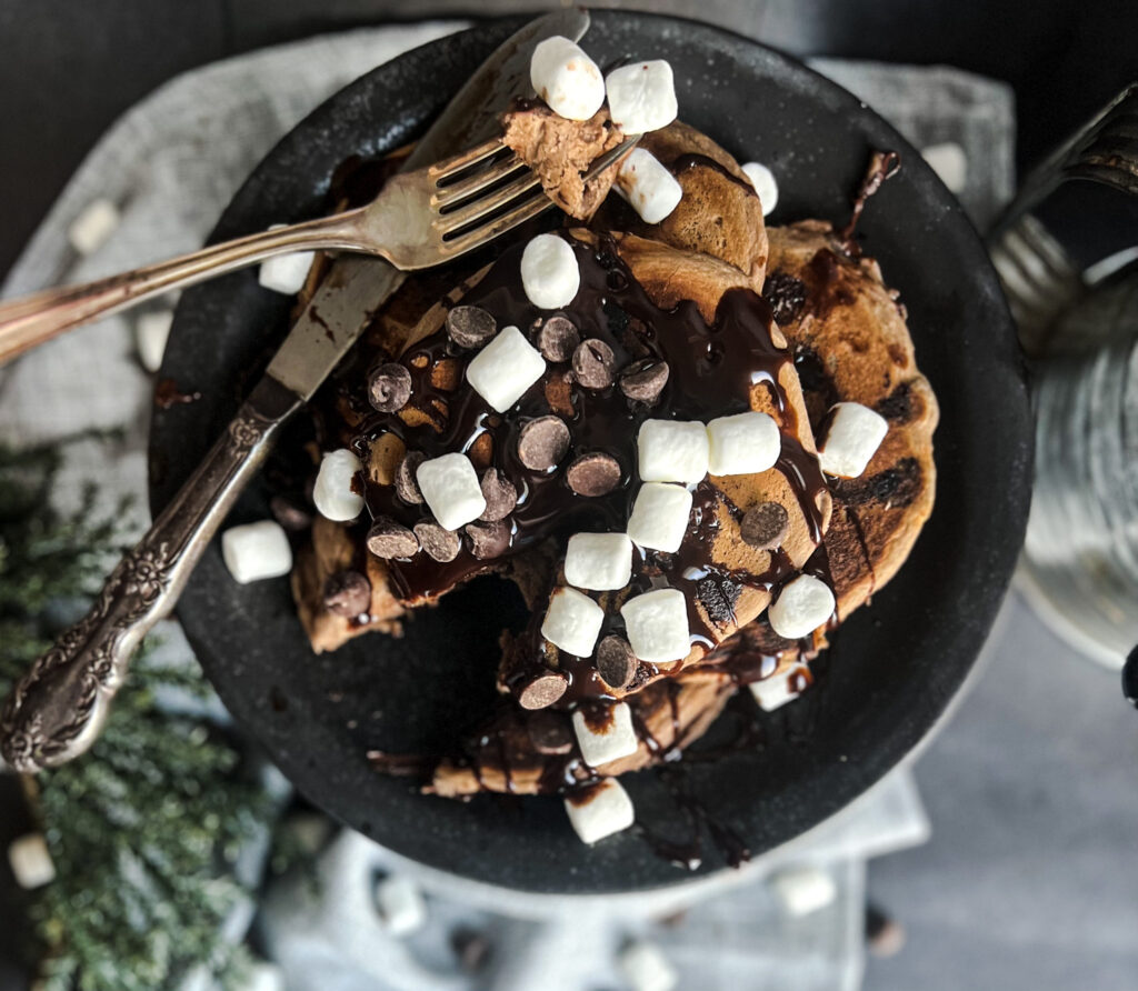 high protein chocolate hot cocoa naked nutrition pancake hot cocoa pancake gluten free dairy free vegan. the pancakes have marshmallows and chocolate sauce on top. The pancakes are sitting on a black plate