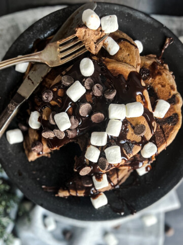 high protein chocolate hot cocoa naked nutrition pancake hot cocoa pancake gluten free dairy free vegan. the pancakes have marshmallows and chocolate sauce on top. The pancakes are sitting on a black plate