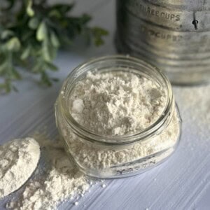 A clear glass jar filled with a light-colored protein pancake mix. A measuring spoon is resting on the mix beside the jar. A flour sifter is in the background.