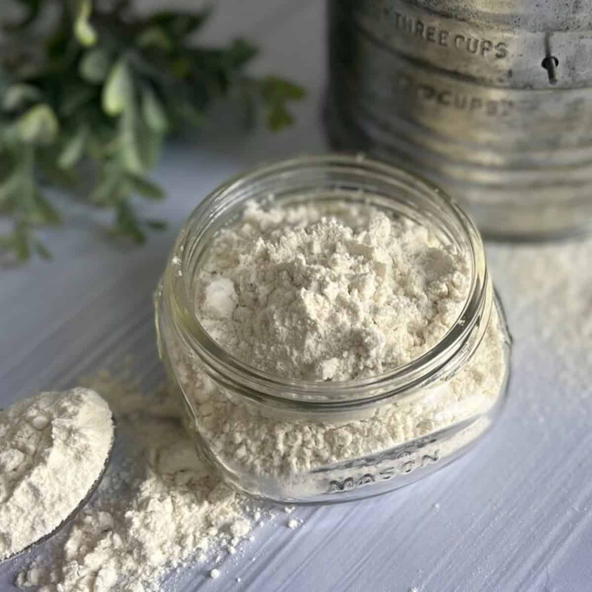 A clear glass jar filled with a light-colored protein pancake mix. A measuring spoon is resting on the mix beside the jar. A flour sifter is in the background.