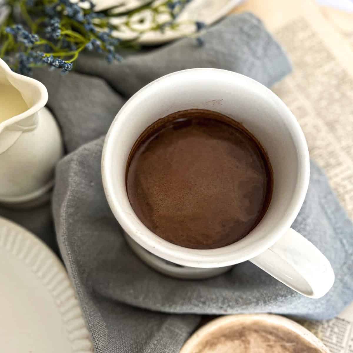 A white mug filled with dark brown, low acid coffee is centered in an overhead shot, resting on a folded gray cloth napkin. A small white creamer pitcher is partially visible to the left.