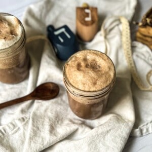 A photo of a jar of chocolate egg cream with a wooden spoon on a towel. The jar is filled with a frothy, chocolate-colored beverage. The wooden spoon is resting on the side of the jar, with a few drops of chocolate egg cream dripping off the end. The background is a neutral color, with a few other kitchen items visible in the distance.