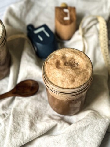 A photo of a jar of chocolate egg cream with a wooden spoon on a towel. The jar is filled with a frothy, chocolate-colored beverage. The wooden spoon is resting on the side of the jar, with a few drops of chocolate egg cream dripping off the end. The background is a neutral color, with a few other kitchen items visible in the distance.