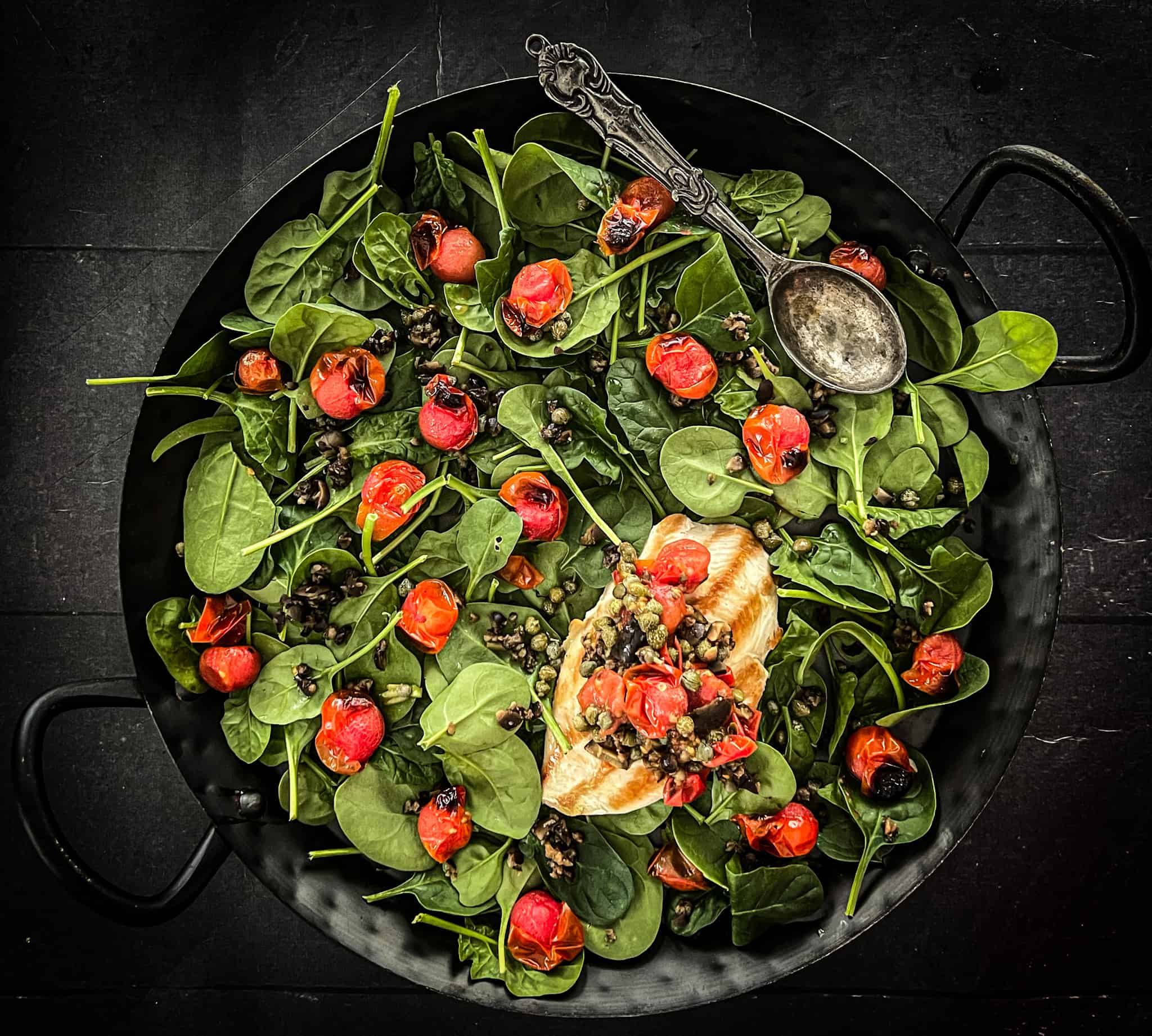 chicken tomatoes capers and spinach arranged in a black cast iron pan