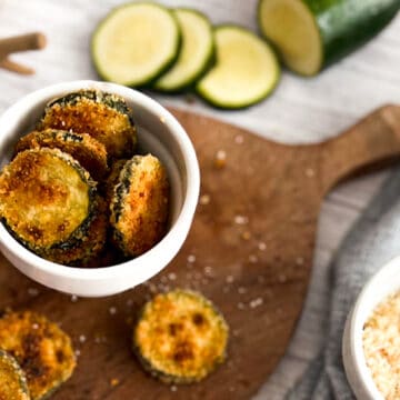 A plate of crispy breaded zucchini chips. The chips are golden brown and have a light dusting of bread crumbs. They are served with a side of ranch dressing.