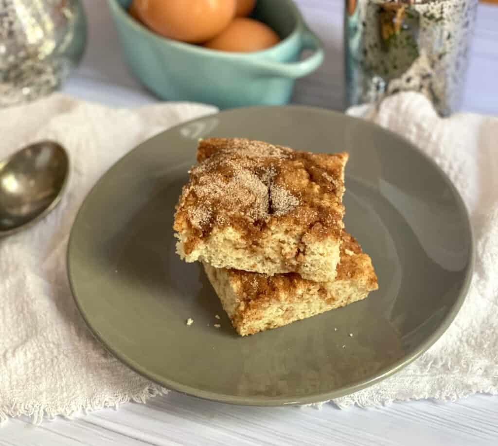 A gray plate has squares of sheet pan breakfast in the middle. The breakfast is made of flour, sugar, eggs and ribbons of cinnamon are swirled thorughout. In the background there is a spoon, eggs and other cooking ingredients. 