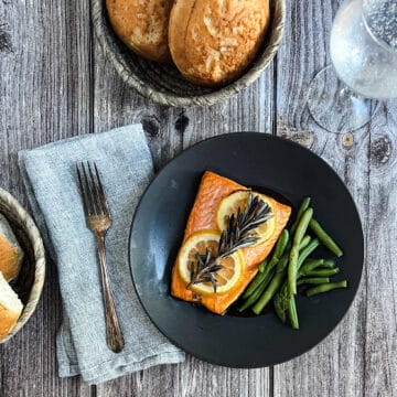The image depicts a beautifully plated dish of "Rosemary Lemon Salmon." The main focus of the image is a generous portion of salmon fillet placed in the center of a black plate. The salmon is pink and topped with lemon slices and a rosemary sprig. Green beans are served as a side and a plate of hot crusty bread is on a small black plate on the side.