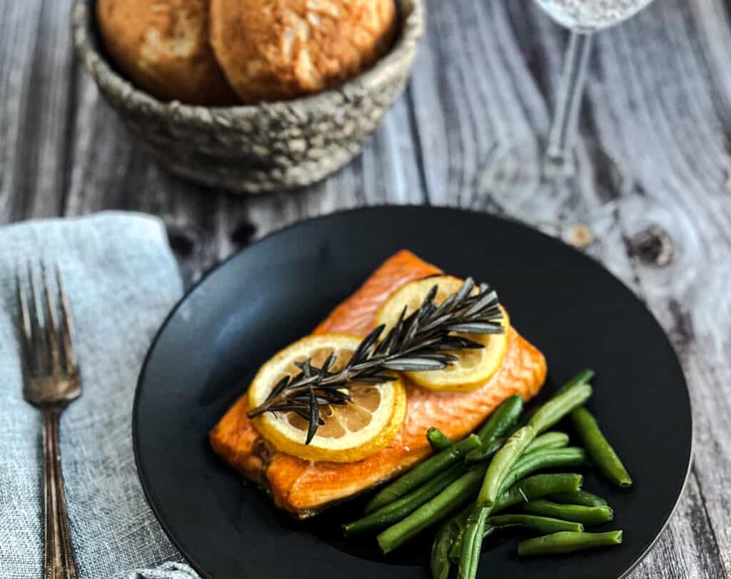 a picture of a fillet of lemon salmon is shown. The salmon is pink and topped with two lemon slices and a fresh sprig of rosemary. Green beans serve as a side dish along with a plate of crusty bread. 
