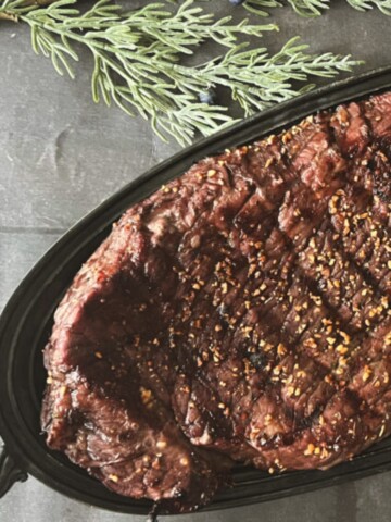 A photo of a London broil steak cooking on a grill. The steak is well-marbled and has a deep brown crust on both sides. It is garnished with a sprig of rosemary and a few sliced cloves of garlic.