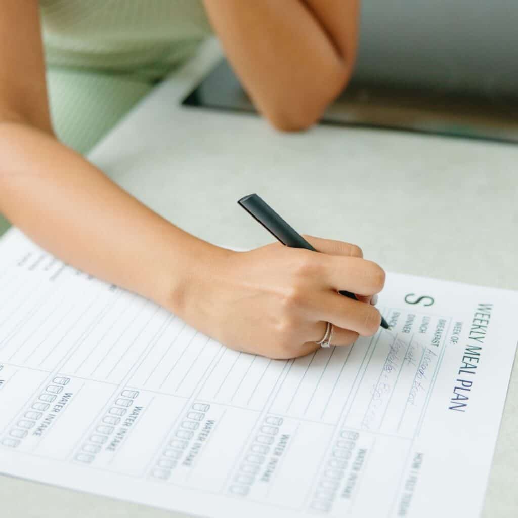 A picture of a person writing a weekly menu. They are holding a black pen in their hand and writing on a paper that has different meals written on it and the words weekly meal plan printed on it. 