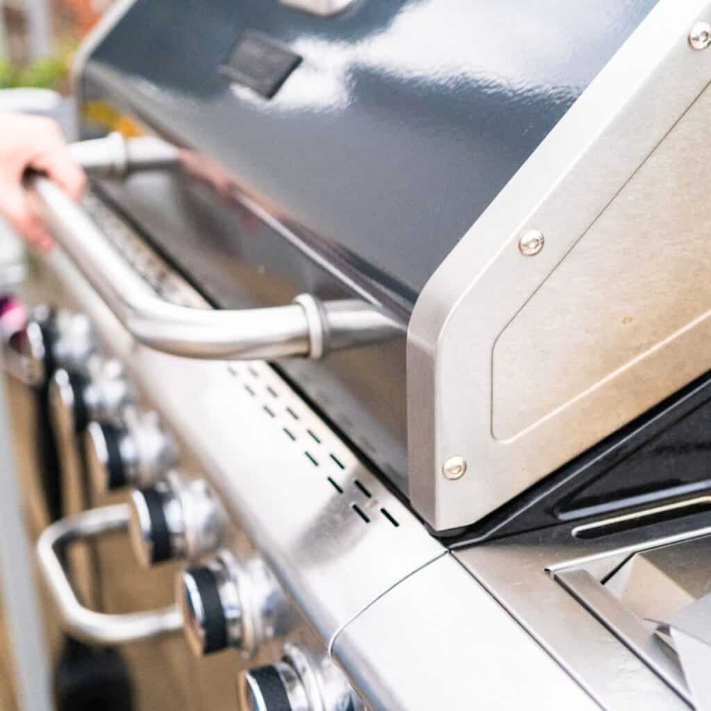 A gas grill smoker with a hinged lid, multiple burners, and a temperature gauge on the front. Wood chips and chunks are placed on the heat source, and a water pan sits on the grill grates