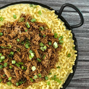 The image shows a skillet filled with macaroni and cheese, pulled pork, and green onions. The skillet is on a wooden table. In the background is a glass of water and a napkin holder. The image is well-lit and the food looks delicious.