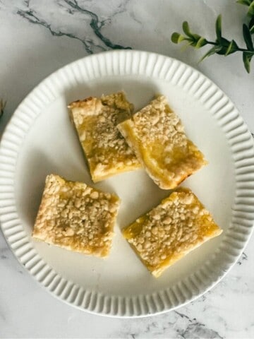 A close-up photo of a lemon bar on a white plate. The lemon bar is square-shaped and has a golden brown crust. The filling is a bright yellow color and has a slightly glossy sheen. The lemon bar is topped with a dusting of powdered sugar.