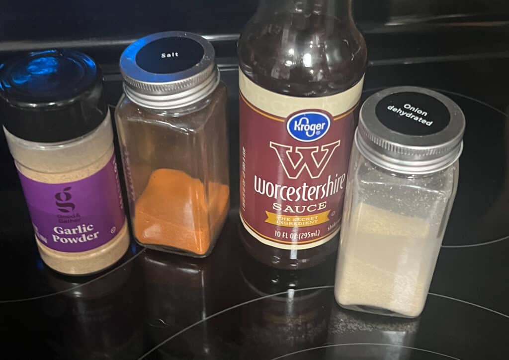 A bottle of Worcestershire sauce sitting on top of a stove, next to a jar of garlic powder and a jar of onion powder and a jar of seasoning salt. 