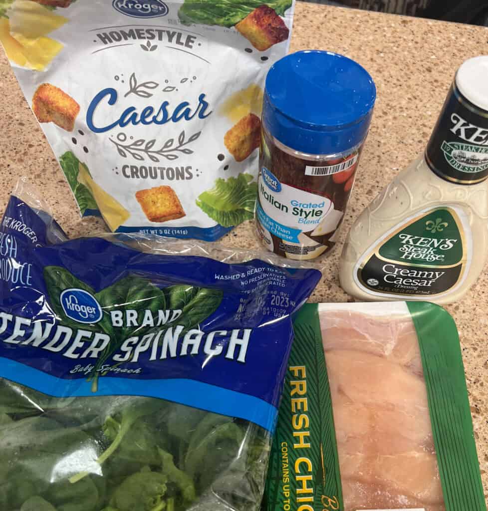 A table with a bag of spinach, a bag of chicken, a bag of croutons, a bottle of caesar dressing, and a bottle of parmesan cheese