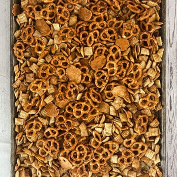 A tray of pretzels, rice chex, corn chex, and bagel chips on a wooden table. There is seasoning added.