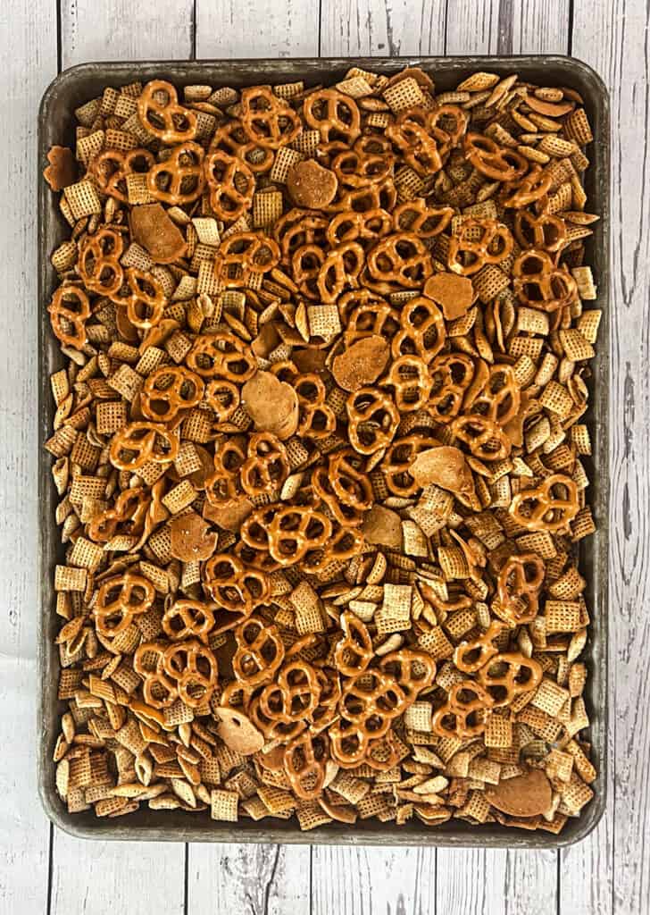 A tray of pretzels, rice chex, corn chex, and bagel chips on a wooden table. There is seasoning added. 