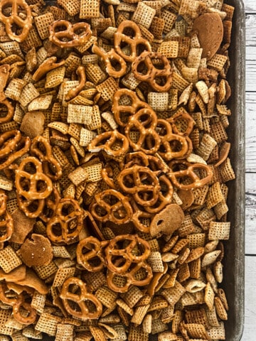 A baking sheet filled with crackers and pretzels, on a wooden table. The mixture also contains bagel chips and seasoning to make homemade chex mix.