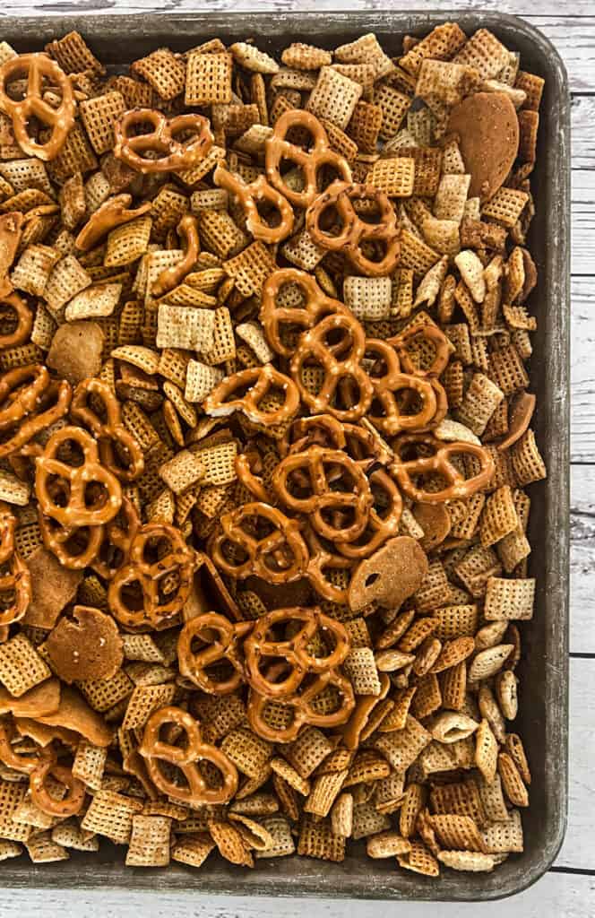 A baking sheet filled with crackers and pretzels, on a wooden table. The mixture also contains bagel chips and seasoning to make homemade chex mix. 
