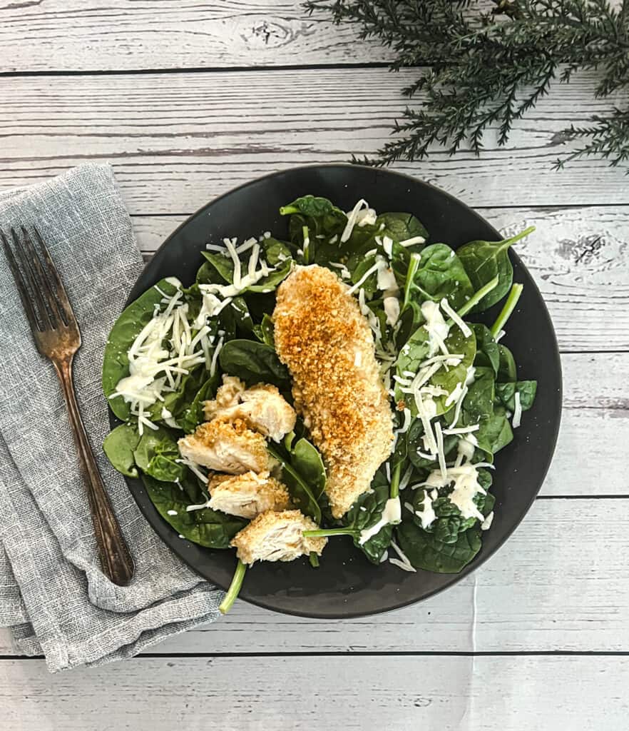 A plate of salad with chicken and spinach on a wooden table. The salad has a variety of greens, including lettuce, spinach, and arugula. There are also chunks of chicken, tomatoes, and cucumbers on the salad. The salad is topped with a creamy caesar dressing